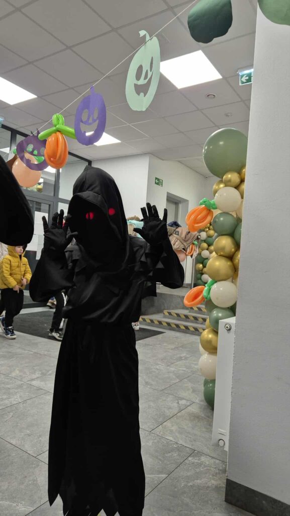 Czarna postać z czerwonymi oczami pozuje w Halloweenowej dekoracji. Tło jest jasne z girlandami dyni. Balony w kolorach złota i zieleni.