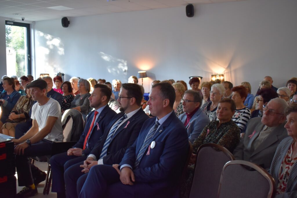 Grupa osób siedzi w jasnym pomieszczeniu. Mężczyźni w garniturach mają różowe wstążki. Ściany są jasnoszare z lampami.
