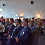 Grupa osób siedzi w jasnym pomieszczeniu. Mężczyźni w garniturach mają różowe wstążki. Ściany są jasnoszare z lampami.