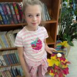 Dziewczynka w różowej koszulce stoi przed półką z książkami. Trzyma kolorowy, jesienny bukiet z liści. W tle widoczne są zielone rośliny.