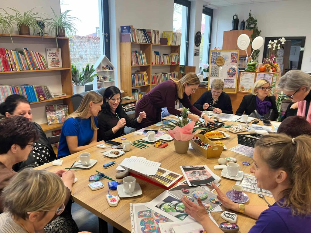 Grupa osób siedzi przy stole z przekąskami i materiałami artystycznymi. W tle regały z książkami i dekoracyjne rośliny. Kolory są jasne z dominacją drewna i zieleni.