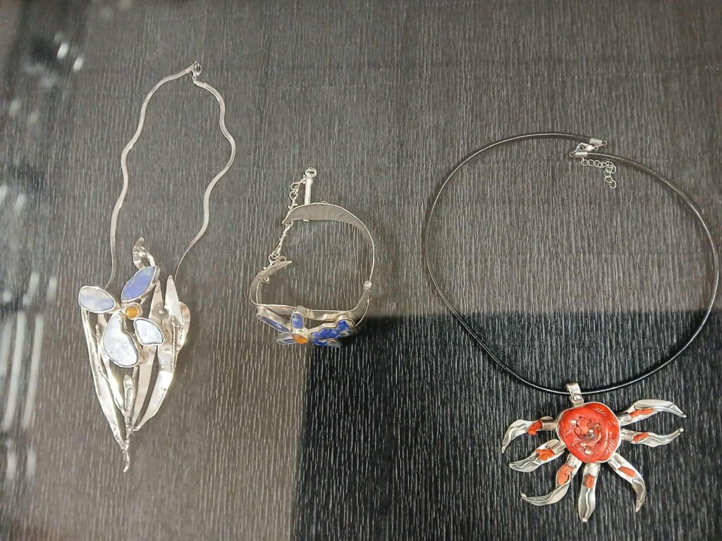 Na zdjęciu widoczne są trzy naszyjniki na ciemnym tle. Dwa mają srebrzyste motywy roślinne z niebieskimi i pomarańczowymi elementami. Trzeci naszyjnik ma czerwony centralny element w kształcie słońca.