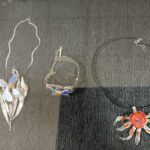 Na zdjęciu widoczne są trzy naszyjniki na ciemnym tle. Dwa mają srebrzyste motywy roślinne z niebieskimi i pomarańczowymi elementami. Trzeci naszyjnik ma czerwony centralny element w kształcie słońca.