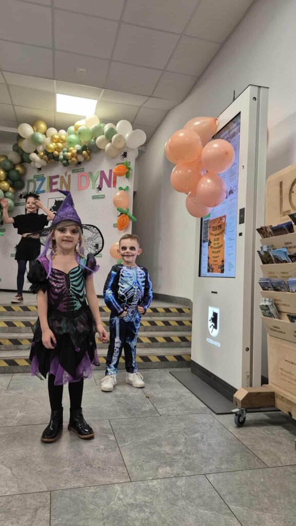 Dwoje dzieci w kostiumach na Halloween, stoją przed dekoracją z balonów. Tło zdobi napis Dzień Dyni i kolorowe balony. Na prawo widoczna jest elektroniczna tablica z informacjami.