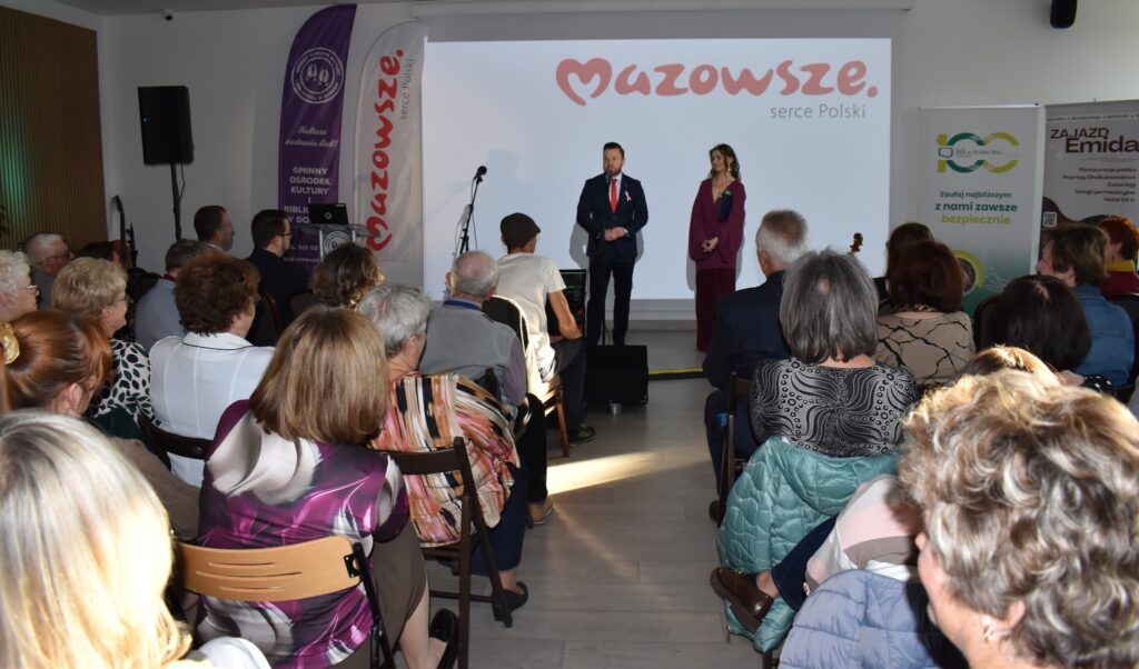 Obraz przedstawia salę z ludźmi, dwoje mówców na scenie, tło z logo Mazowsza. Dominuje jasne oświetlenie i ciepłe kolory.