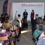 Obraz przedstawia salę z ludźmi, dwoje mówców na scenie, tło z logo Mazowsza. Dominuje jasne oświetlenie i ciepłe kolory.