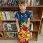 Dziecko w niebieskiej koszulce trzyma kolorowy stroik z liści. W tle brązowy regał z różnorodnymi książkami.