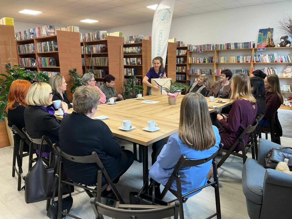 Na zdjęciu widać grupę osób siedzących przy stole w bibliotece. W tle półki z książkami, na stole filiżanki. Osoba stojąca prowadzi dyskusję.