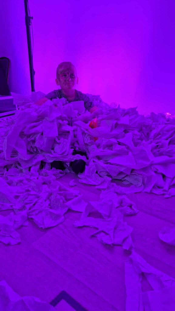 Na zdjęciu jest osoba otoczona mnóstwem papieru. Oświetlenie w kolorze fioletowym dominuje cały obraz. Tło jest jednolite, co podkreśla centralną scenę.