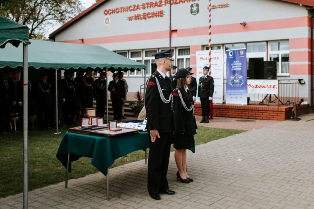 Ceremonia przed strażnicą OSP w Mileńcu, ludzie w mundurach. Na pierwszym planie stół z nagrodami, w tle namiot i budynek.