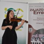 Kobieta gra na skrzypcach, stojąc przed dwoma banerami. Ubrana jest na czarno, tło jest jasne z tekstem i grafiką. Na jednym banerze widoczna jest reklama hotelu.