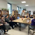 Na zdjęciu widać grupę osób siedzących przy stole w bibliotece. Półki z książkami i rośliny dekorują przestrzeń, a dominują kolory ziemiste i drewno. Osoba w fioletowej koszulce prezentuje coś na biurku.