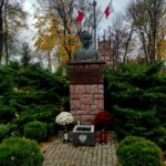 Popiersie na kamiennym postumencie w parku z flagami Polski w tle. Zielone krzewy i jesienne drzewa otaczają pomnik. Kwiaty i znicze u podstawy.