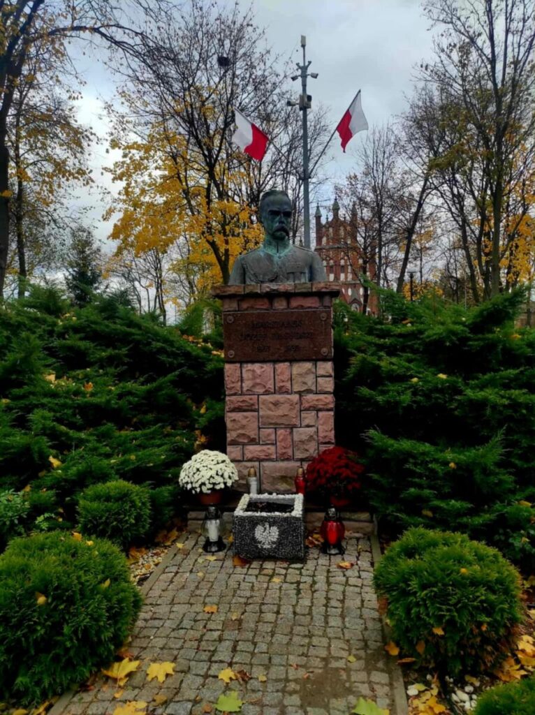 Popiersie na kamiennym postumencie w parku z flagami Polski w tle. Zielone krzewy i jesienne drzewa otaczają pomnik. Kwiaty i znicze u podstawy.