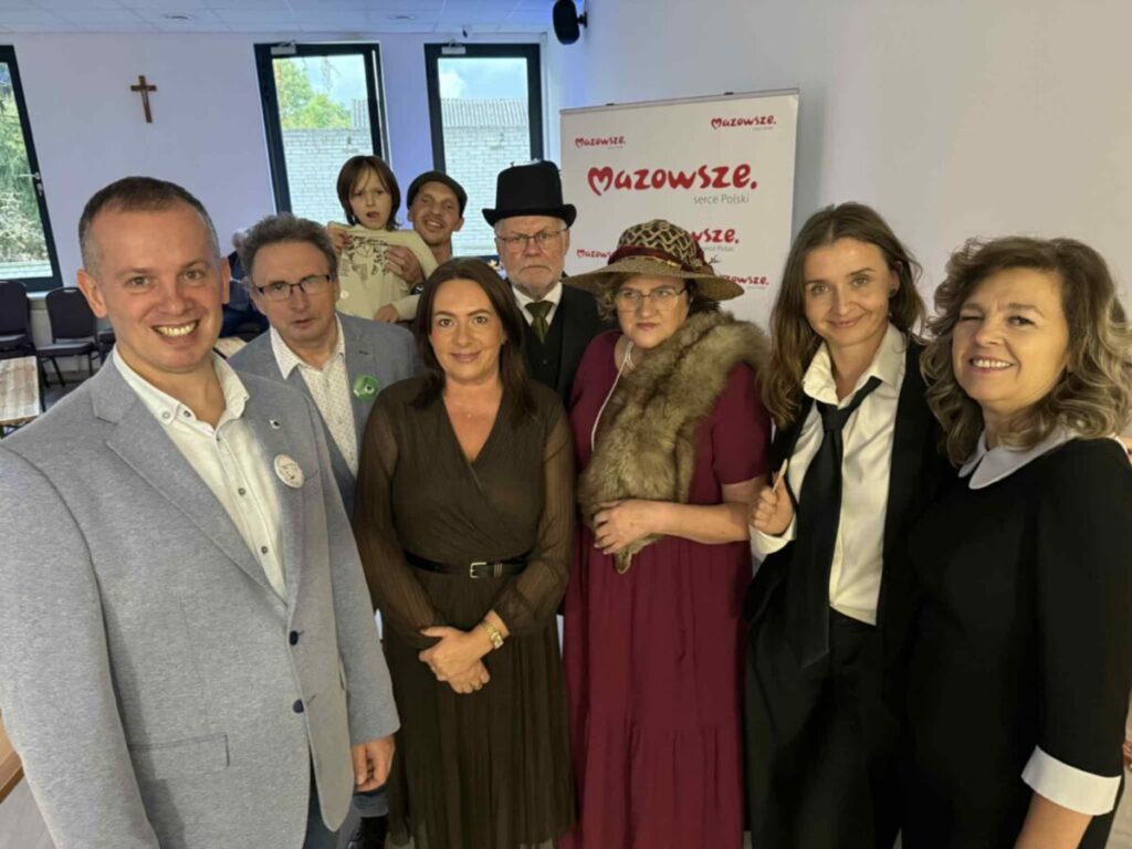 Grupa siedmiu dorosłych i jedno dziecko pozuje na tle ścianki z napisem “Mazowsze”, pokój jasny z dużymi oknami.