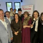 Grupa siedmiu dorosłych i jedno dziecko pozuje na tle ścianki z napisem “Mazowsze”, pokój jasny z dużymi oknami.