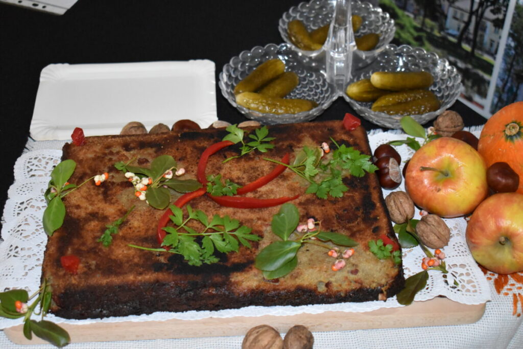 Kwadratowy, brązowy placek z ziołami i papryką na białej serwetce. Obok jabłka, orzechy i ogórki na czarnym tle. Ovalne miski na ogórki.