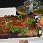 Kwadratowy, brązowy placek z ziołami i papryką na białej serwetce. Obok jabłka, orzechy i ogórki na czarnym tle. Ovalne miski na ogórki.