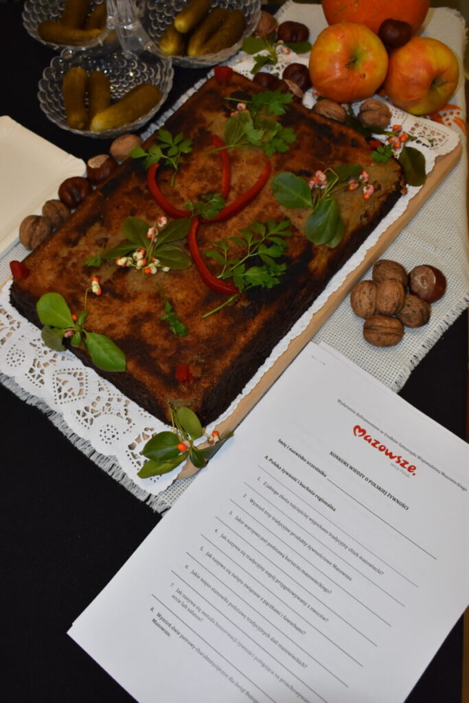 Na białym obrusie leży prostokątne ciasto ozdobione ziołami i warzywami. Obok są orzechy, jabłka i miska z ogórkami. W prawym dolnym rogu widoczny jest dokument z logo Mazowsze.