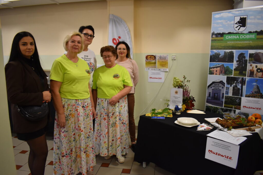 Pięć osób stoi przy stole z jedzeniem i materiałami promocyjnymi. Dwie osoby mają zielone koszulki i kolorowe spódnice. Tło jest jasne, z banerem Gmina Dobre.