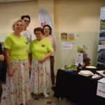 Pięć osób stoi przy stole z jedzeniem i materiałami promocyjnymi. Dwie osoby mają zielone koszulki i kolorowe spódnice. Tło jest jasne, z banerem Gmina Dobre.