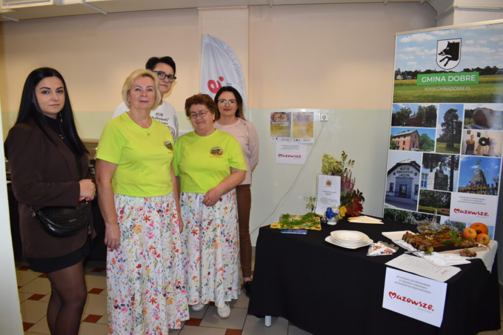 Pięć osób stoi obok stoiska z ulotkami i jedzeniem. Dwie kobiety w zielonych koszulkach mają kwieciste spódnice. Tło jasne, z plakatem promującym Gminę Dobre.