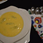 Talerz z żółtą zupą na białym talerzu, obok kolorowa serwetka z łyżkami na ciemnym tle.