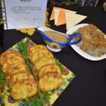 Na stole leżą placki ziemniaczane i sos w kolorowej sosjerce. W tle widać serwetki i tabliczkę z napisem. Dominują kolory żółty i zielony.