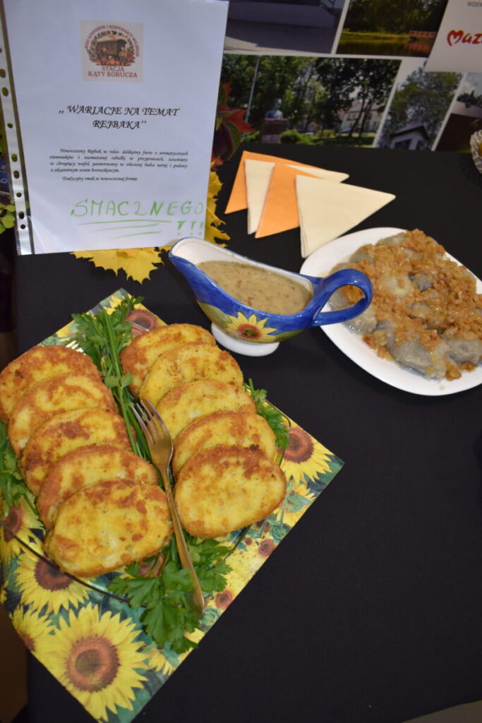 Na stole leżą placki ziemniaczane i sos w kolorowej sosjerce. W tle widać serwetki i tabliczkę z napisem. Dominują kolory żółty i zielony.