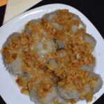 Na białym talerzu leżą szare kluski z brązową skwarkową posypką. Tło jest czarne z żółtą serwetką. Danie jest obfite i domowe.