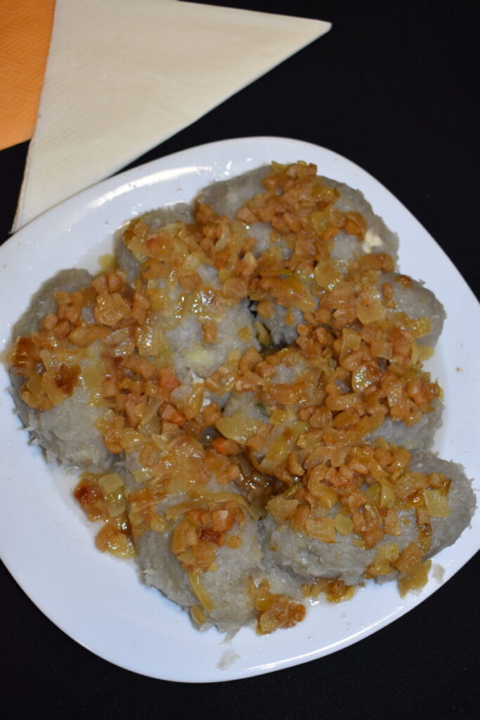 Na białym talerzu leżą szare kluski z brązową skwarkową posypką. Tło jest czarne z żółtą serwetką. Danie jest obfite i domowe.