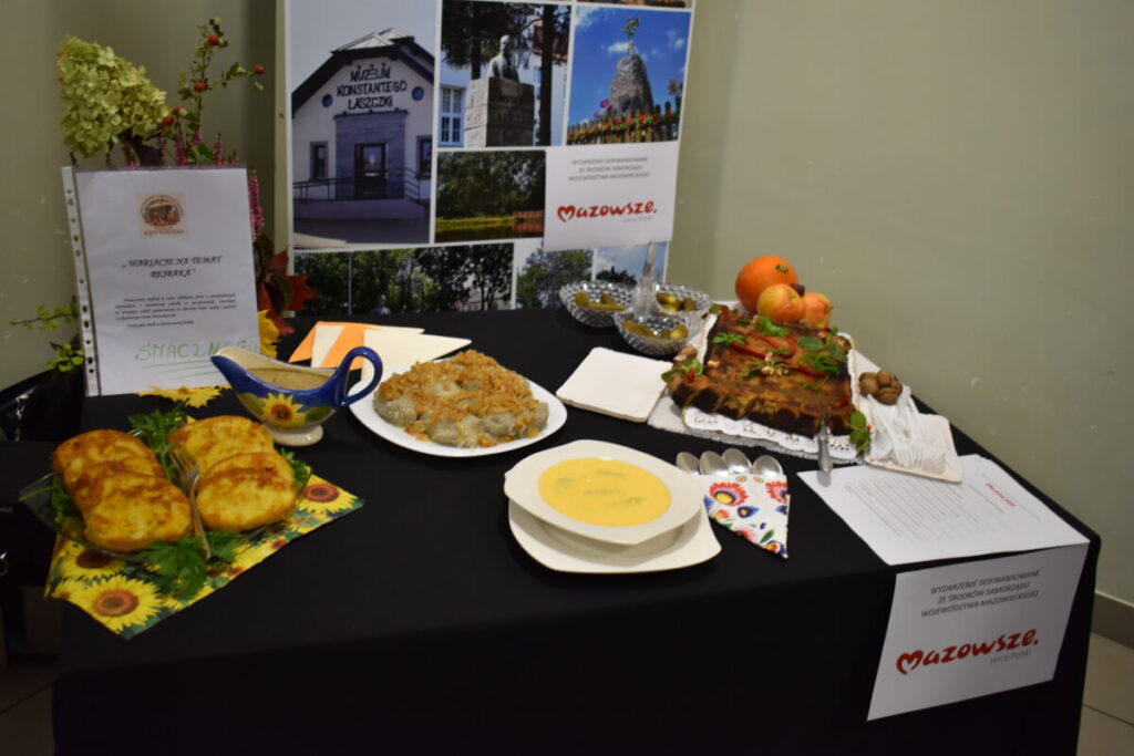 Stół z tradycyjnymi potrawami, w tym zupa, placki i ciasto, na czarnym obrusie. Tło w neutralnym kolorze z plakatem muzeum. Kolorowe serwetki i ozdobne naczynia.