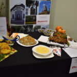 Stół z tradycyjnymi potrawami, w tym zupa, placki i ciasto, na czarnym obrusie. Tło w neutralnym kolorze z plakatem muzeum. Kolorowe serwetki i ozdobne naczynia.