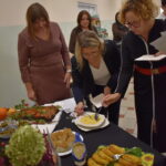 Grupa osób przy stole degustuje dania. Na stole kolorowe potrawy, zupy i przekąski. Jasne tło z kremowymi ścianami.