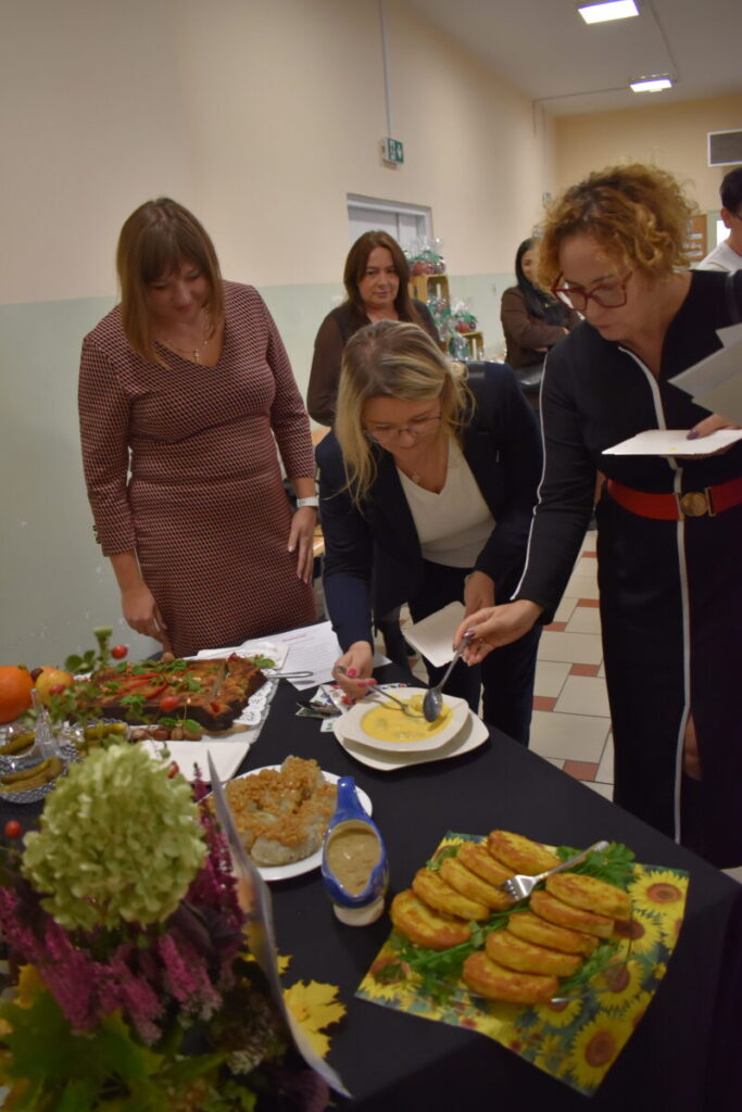 Grupa osób przy stole degustuje dania. Na stole kolorowe potrawy, zupy i przekąski. Jasne tło z kremowymi ścianami.