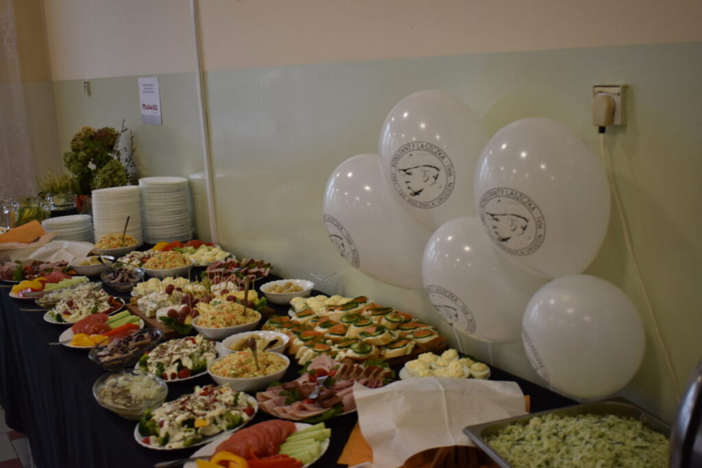 Na stole z ciemnym obrusem ułożono różnorodne sałatki i kanapki. Obok stoją białe balony z nadrukiem oraz stos talerzy. Tło jest jasne, dekoracje są minimalistyczne.