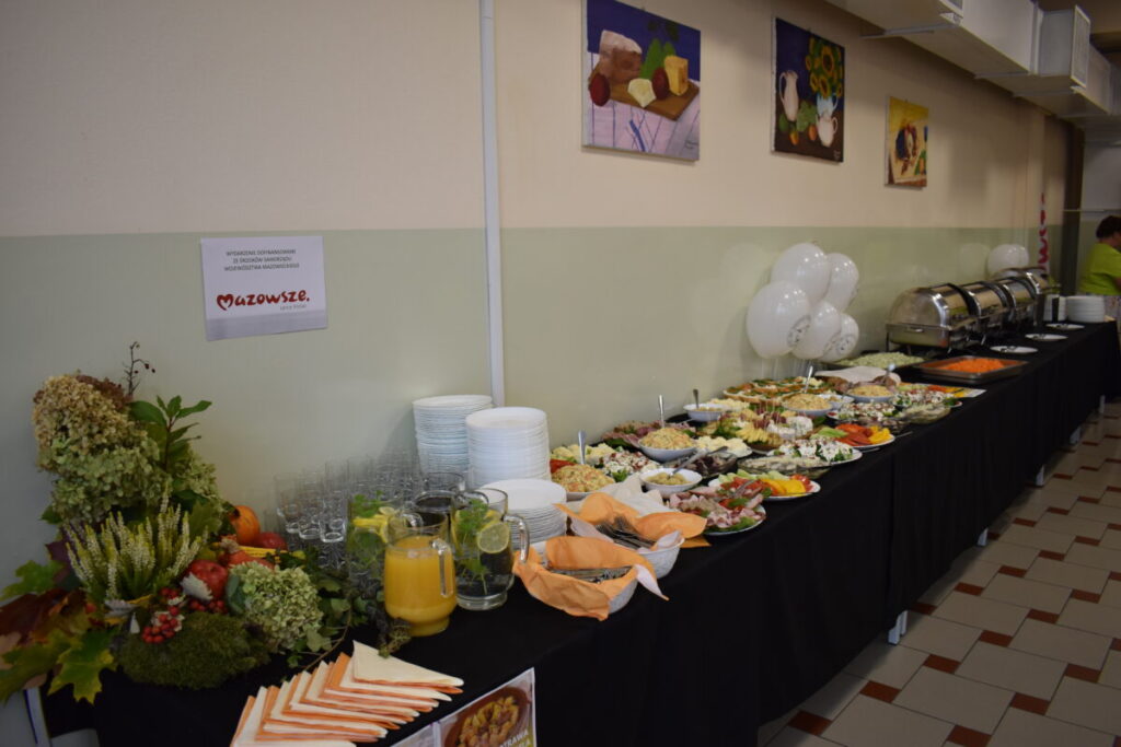 Stół szwedzki z daniami na czarnym obrusie. Tło beżowe z obrazami, balony i zastawa. Posiłki w kolorach warzyw i owoców.