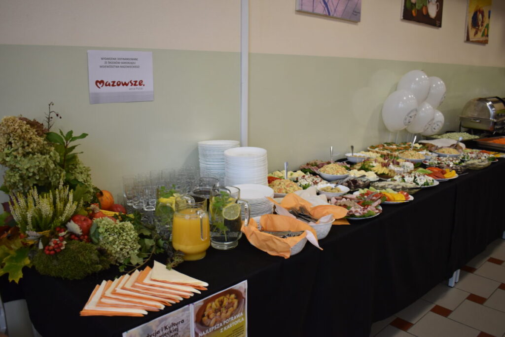 Stół zastawiony różnorodnymi potrawami na białych talerzach, obok bukiet kwiatów. Tło jasne, ściana zielonkawa. Po lewej stronie stoi dzbanek z sokiem pomarańczowym.