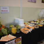 Stół zastawiony różnorodnymi potrawami na białych talerzach, obok bukiet kwiatów. Tło jasne, ściana zielonkawa. Po lewej stronie stoi dzbanek z sokiem pomarańczowym.