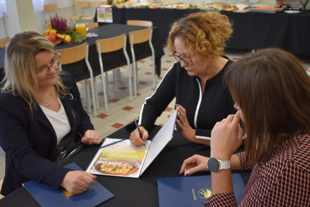 Trzy kobiety siedzą przy stole i przeglądają dokumenty. Tło wypełniają krzesła i stół z czarnym obrusem. Kolory: czarny, biały, jasnobrązowy.