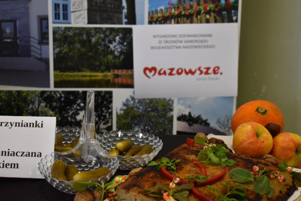 Stół z potrawami regionalnymi: ziemniaki, kiszone ogórki, ozdoby z ziół. W tle plansza Mazowsze. Jabłka i dynia obok.