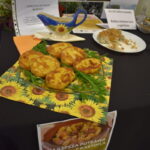 Na czarnym stole leży talerz z plackami ziemniaczanymi na serwetce w słoneczniki. Obok dekoracyjny dzbanek i biała tabliczka. W tle widoczny plakat z nagrodą za najlepsze danie z kartofla.