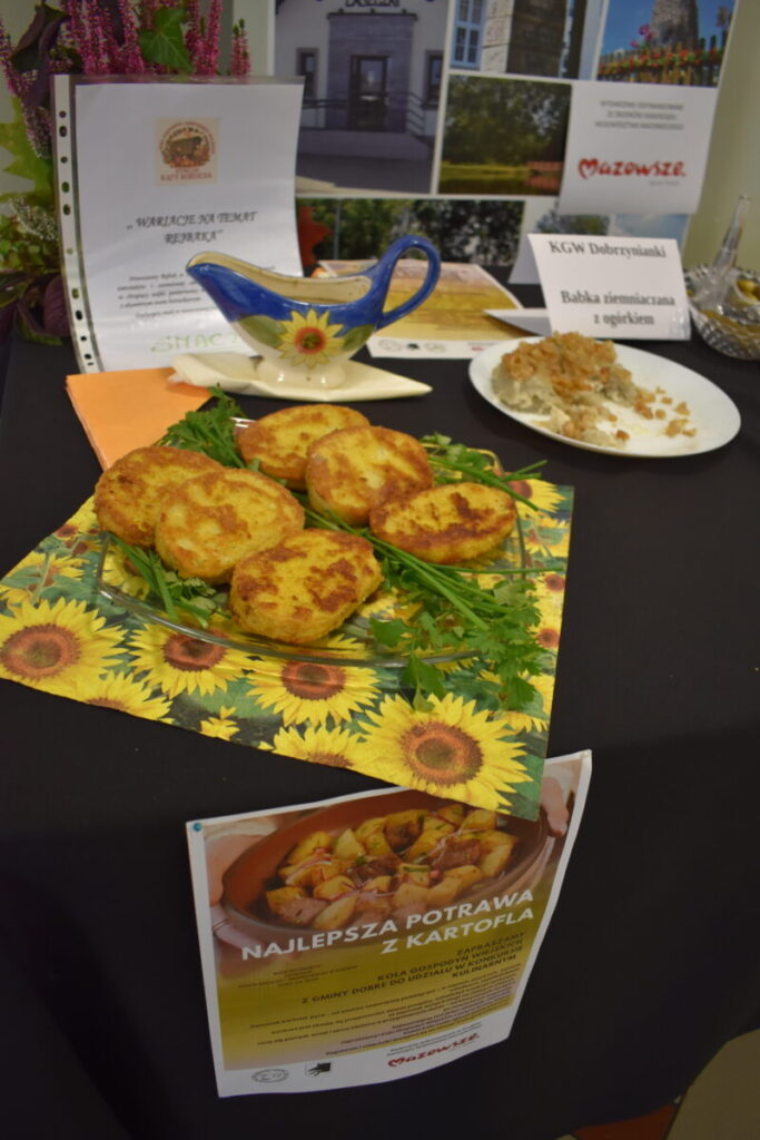 Na czarnym stole leży talerz z plackami ziemniaczanymi na serwetce w słoneczniki. Obok dekoracyjny dzbanek i biała tabliczka. W tle widoczny plakat z nagrodą za najlepsze danie z kartofla.