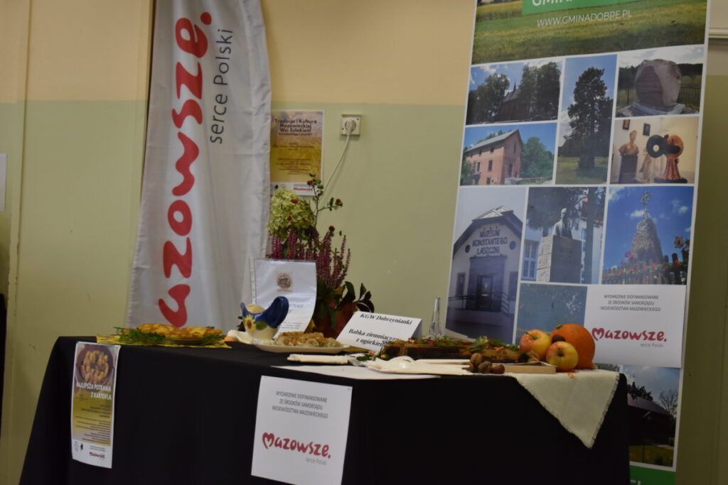Stoisko z regionalnymi potrawami z Mazowsza, z dekoracjami w postaci owoców i kwiatów. Tło jasne z plakatami promującymi region. Dominują kolory: biały, czerwony i zielony.