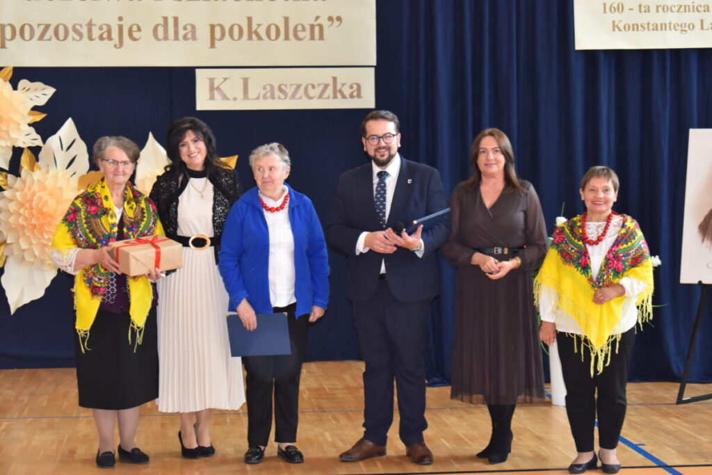 Sześć osób stoi przed ciemnoniebieskim tłem. Dwie kobiety mają kolorowe chusty. Na górze znajduje się napis „K. Laszczka.”