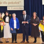 Sześć osób stoi przed ciemnoniebieskim tłem. Dwie kobiety mają kolorowe chusty. Na górze znajduje się napis „K. Laszczka.”