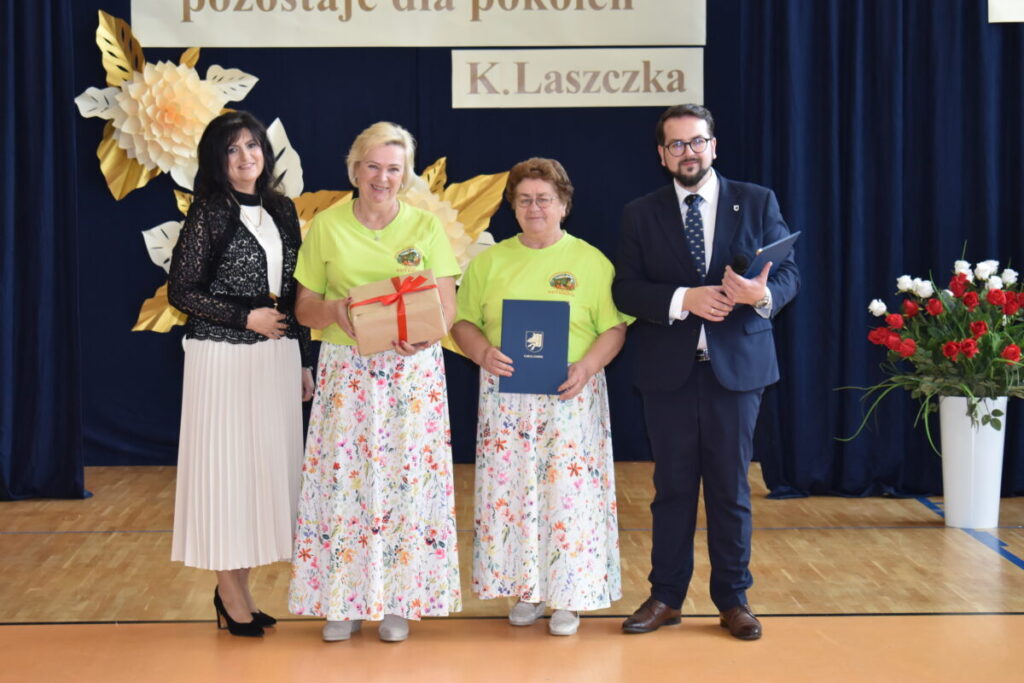 Cztery osoby stoją na tle ciemnoniebieskiej zasłony z dekoracją z papierowych kwiatów. Dwie kobiety w zielonych koszulkach i spódnicach w kwiaty trzymają nagrody. Obok stoją elegancko ubrani kobieta i mężczyzna.