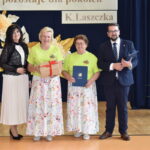 Cztery osoby stoją na tle ciemnoniebieskiej zasłony z dekoracją z papierowych kwiatów. Dwie kobiety w zielonych koszulkach i spódnicach w kwiaty trzymają nagrody. Obok stoją elegancko ubrani kobieta i mężczyzna.