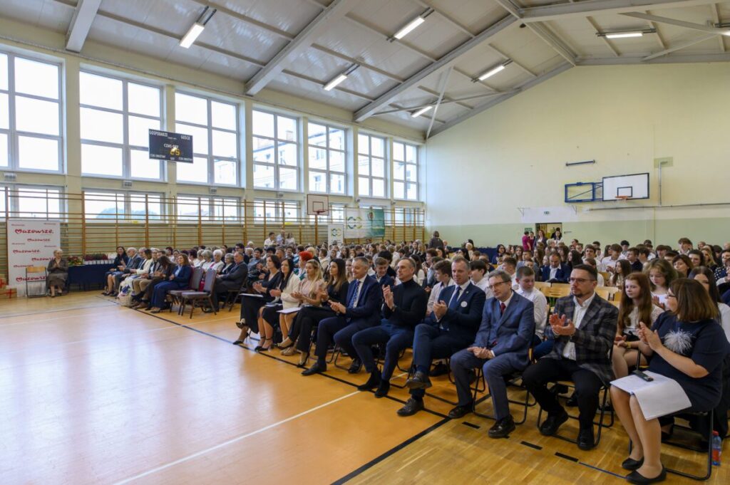 Sala gimnastyczna z jasnym ścianami i oknami, wypełniona ludźmi na krzesłach. Na podłodze jasne panele, kosz do koszykówki w tle. Wszyscy obecni siedzą i patrzą w jednym kierunku.