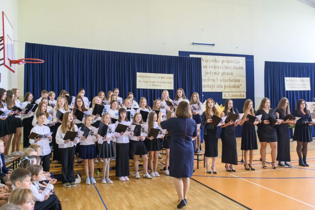 Grupa dzieci i dorosłych śpiewa na sali gimnastycznej. Ubrani w białe i czarne stroje, stoją przed niebieską kurtyną. Na ścianie widoczne są plakaty z napisami.
